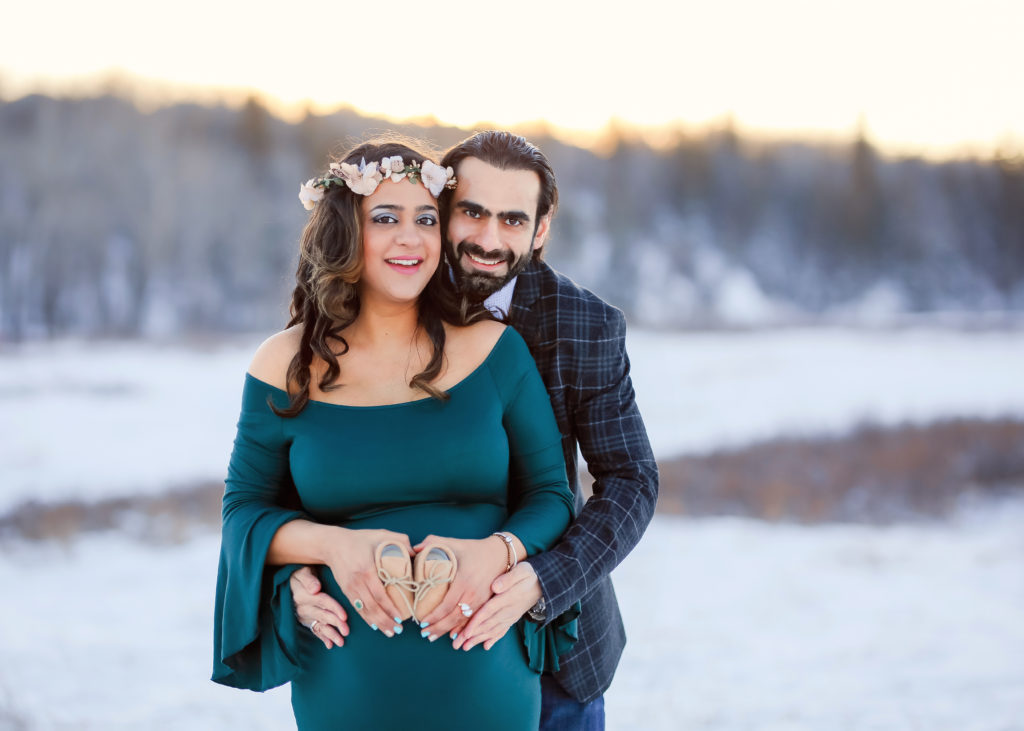 alt="mother to be in a long hunter green dress with husband behind her arms wrapped around her and they are holding baby shoes together smiling at the camera in the frosty winter month of January at sunset"
