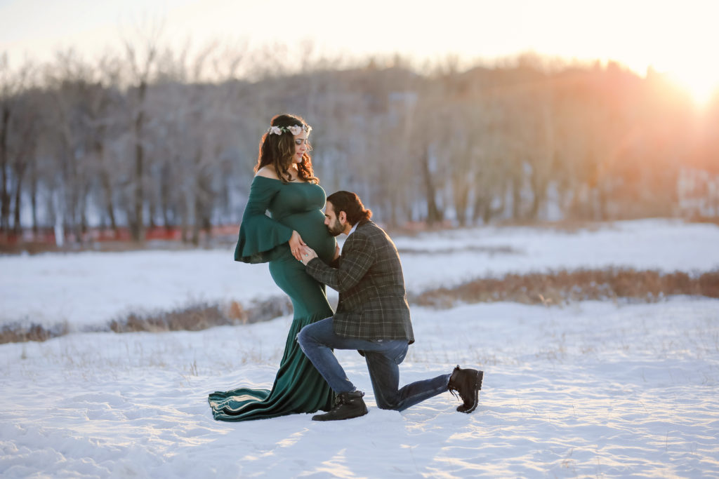 alt="mother to be in a long hunter green dress with husband on one knee holding and kissing her baby belly in the frosty winter month of January at sunset"
