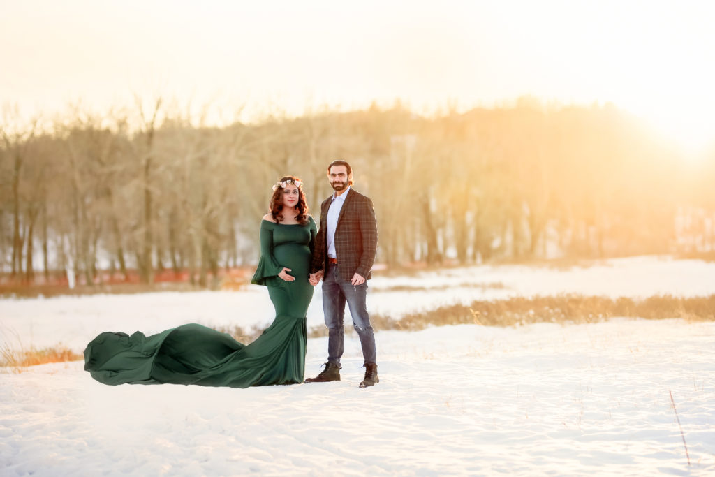 alt="mother to be in a long hunter green dress holding her husbands hand in the frosty winter month of January at sunset"