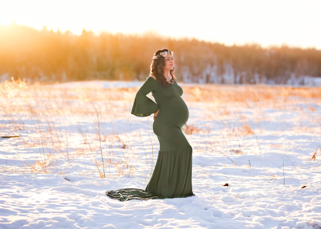 alt="mother to be in a long hunter green dress in the frosty winter month of January at sunset wearing a floral crown"