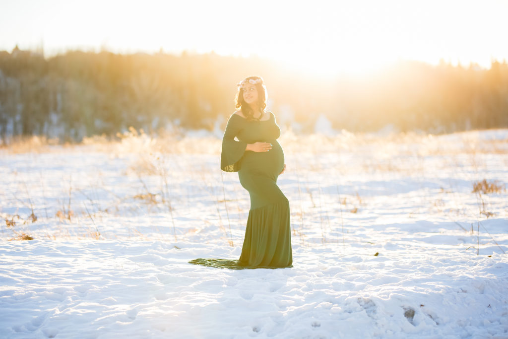 alt="mother to be in a long hunter green dress with her head looking over her shoulder in the frosty winter month of January at sunset wearing a floral crown"