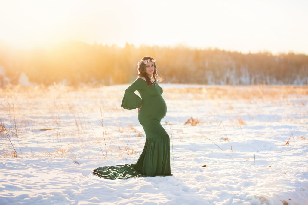 alt="mother to be in a long hunter green dress looking at the camera smiling in the frosty winter month of January at sunset wearing a floral crown"