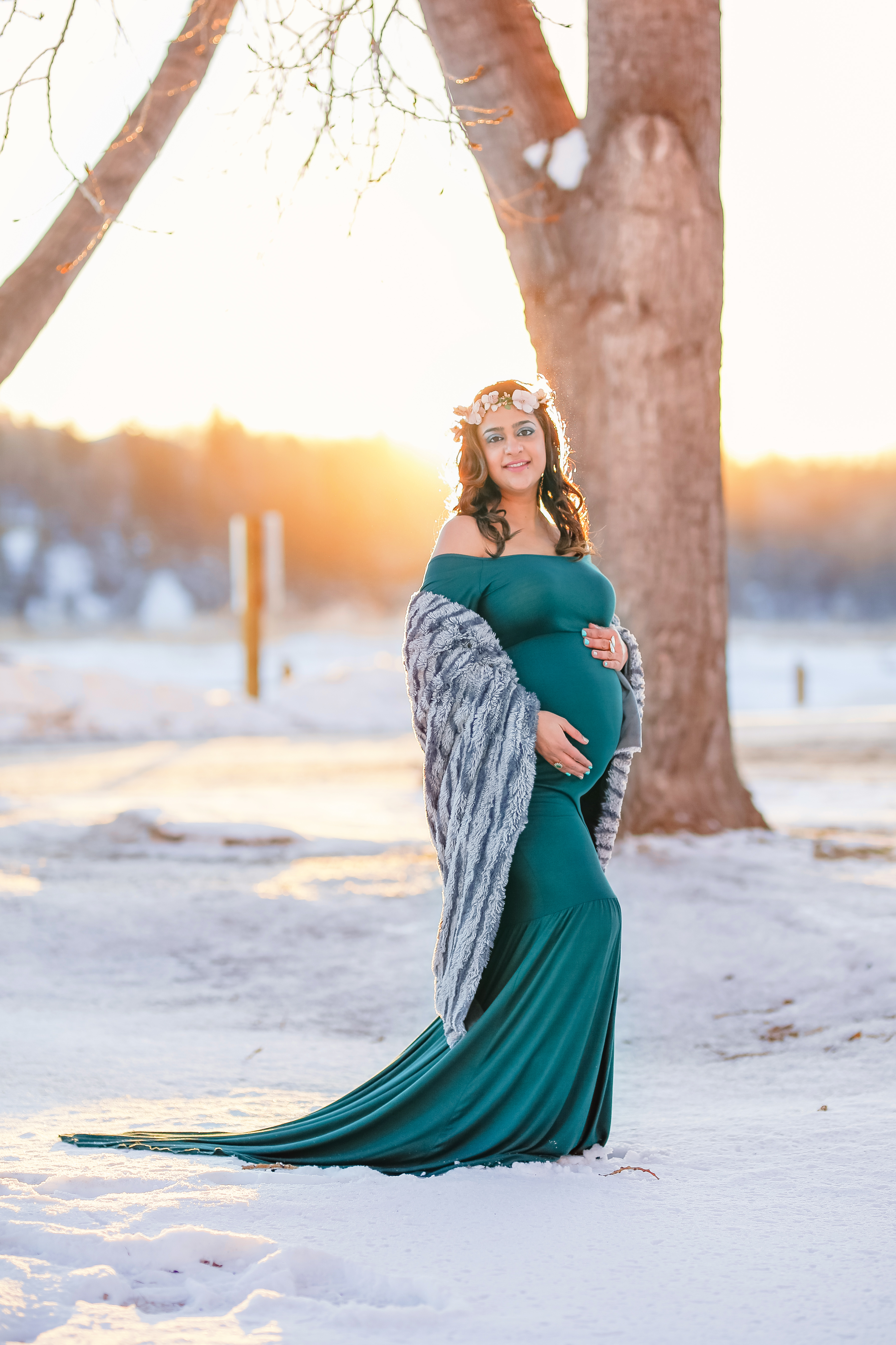 alt="mother to be in a long hunter green dress and a fur wrap around her arms with the sun behind her in the frosty winter month of January at sunset"