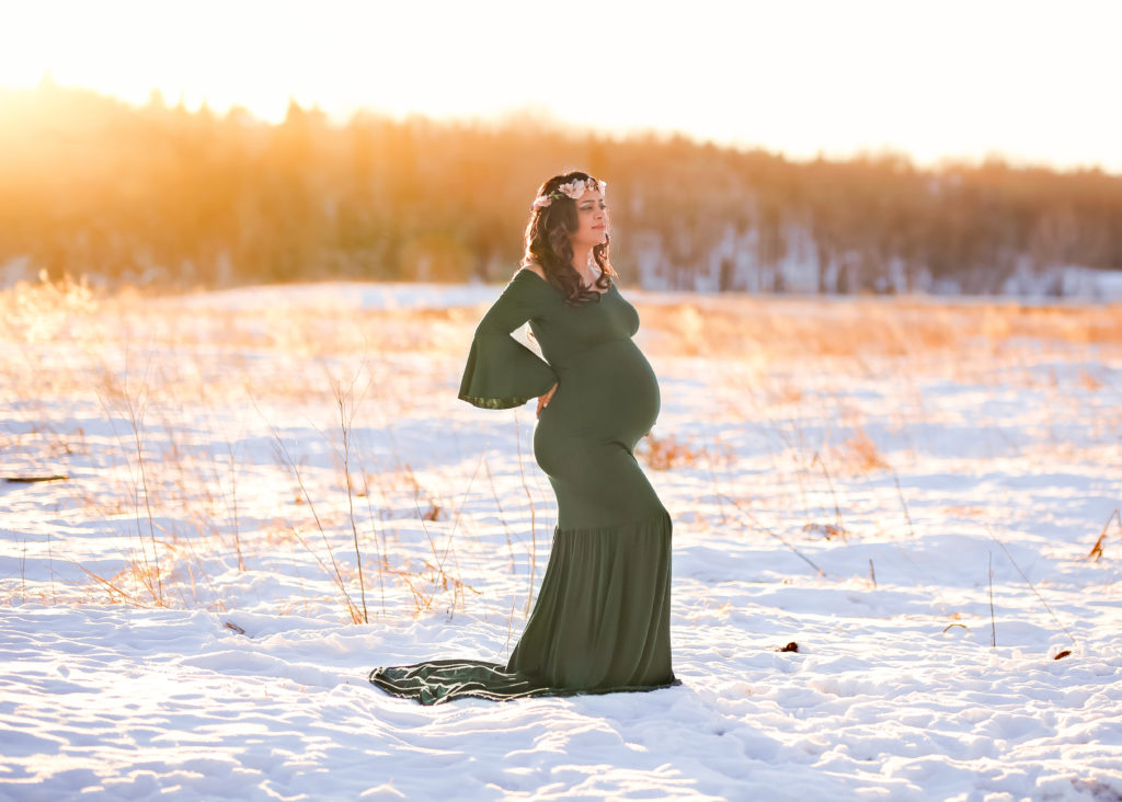alt="mother to be in a long hunter green dress with her hand on the lower back in the frosty winter month of January at sunset wearing a floral crown"