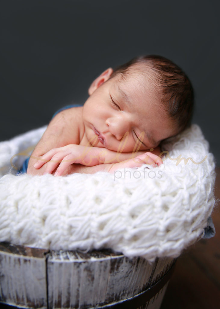 alt="newborn baby boy wrapped in a blue in a bucket with chin on hands pose"
