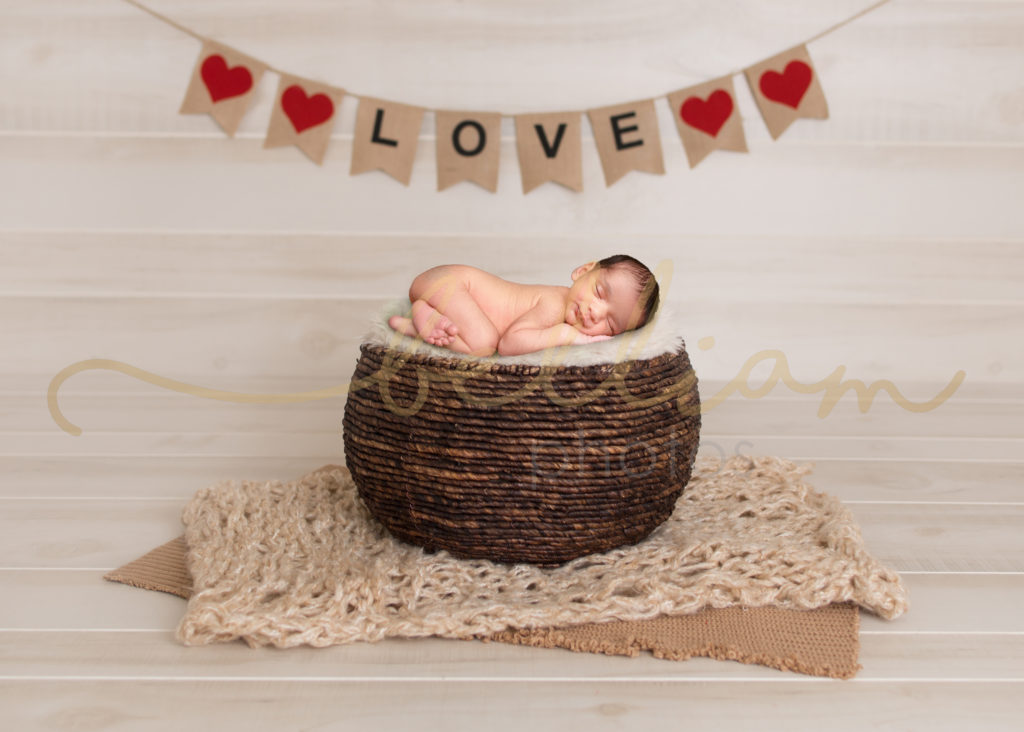 alt="newborn baby boy in bum up pose in a basket with a love sign above"