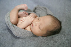 alt="newborn baby boy in a grey wrap curled up side view"