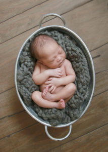 alt="newborn baby boy in a grey wrap curled up in a bucket on a wood floor"