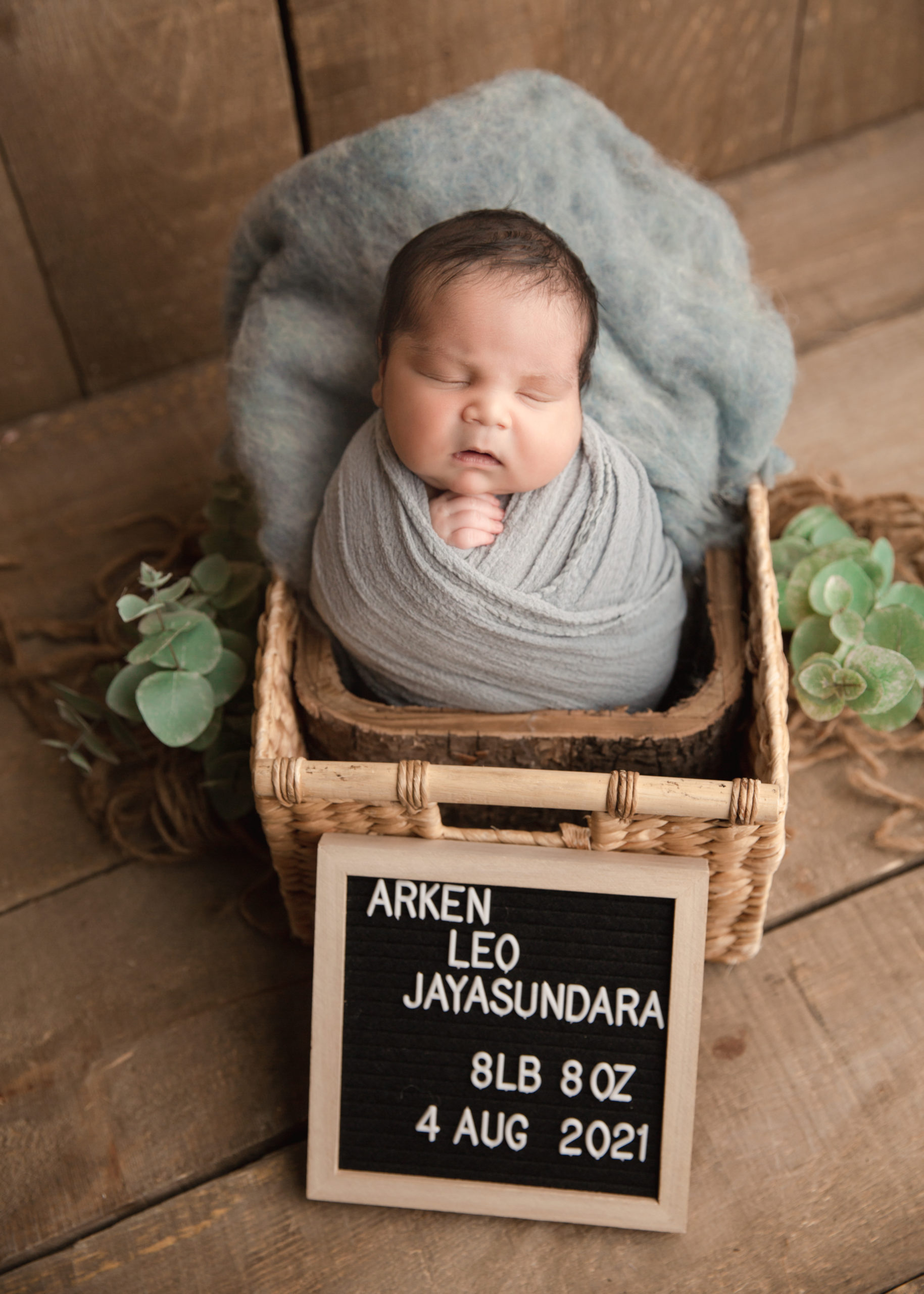 baby boy in a basket wrapped in a blue wrap with name board
