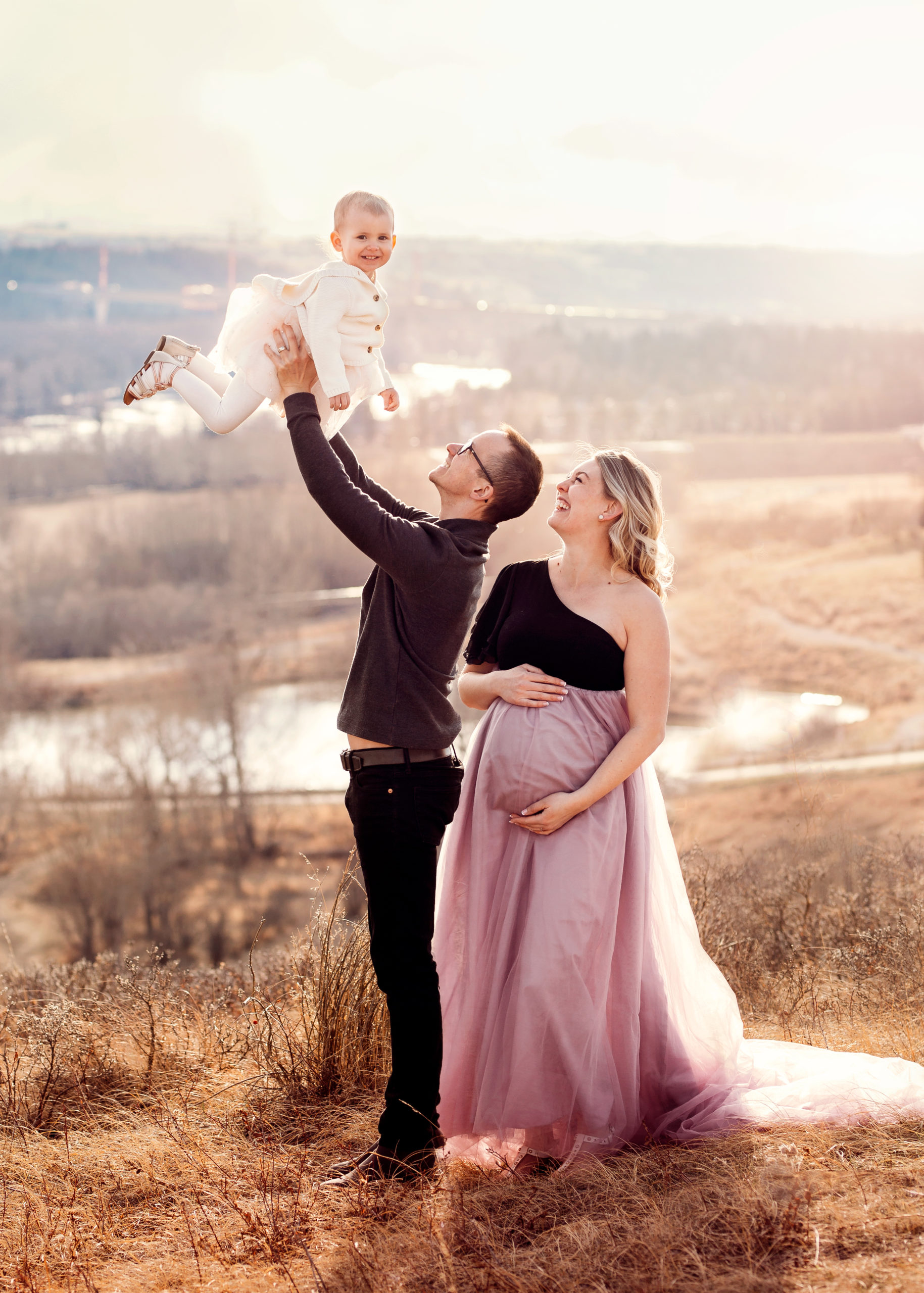 Young couple holding thier toddler in the air while mom holds her pregnant belly